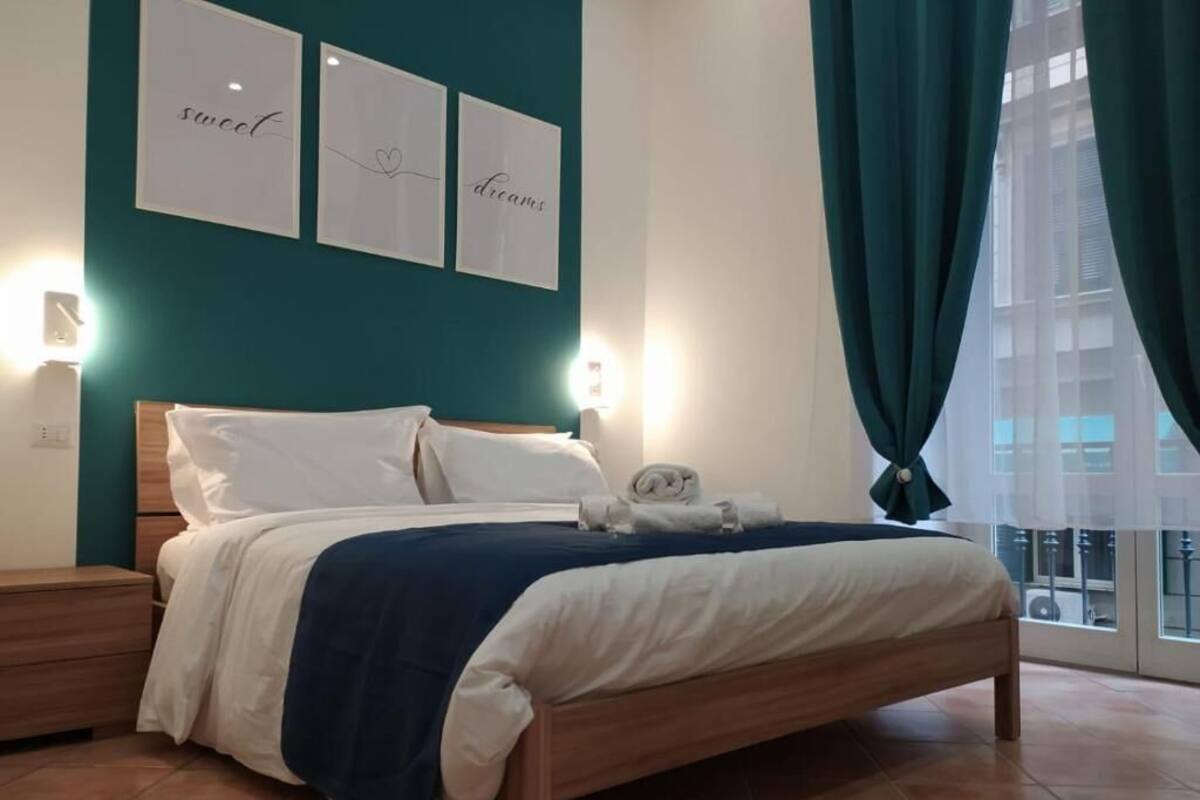 Un lit bien fait, avec une literie blanche et bleue, une tête de lit en bois et une table de chevet avec une lampe. Deux tableaux encadrés sont accrochés au mur, l’un avec le mot « doux » et l’autre avec un symbole en forme de cœur.