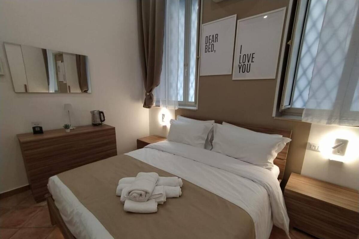 Un lit bien fait, avec des serviettes pliées, une commode en bois, une table de chevet avec une lampe, et deux panneaux encadrés accrochés au mur.