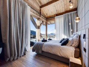 A bedroom with a large bed, wooden flooring, and a view of mountains through the window. - Luxury chalet with wellness - 14 pers, sauna, fireplace (Montvalezan-La Rosière)