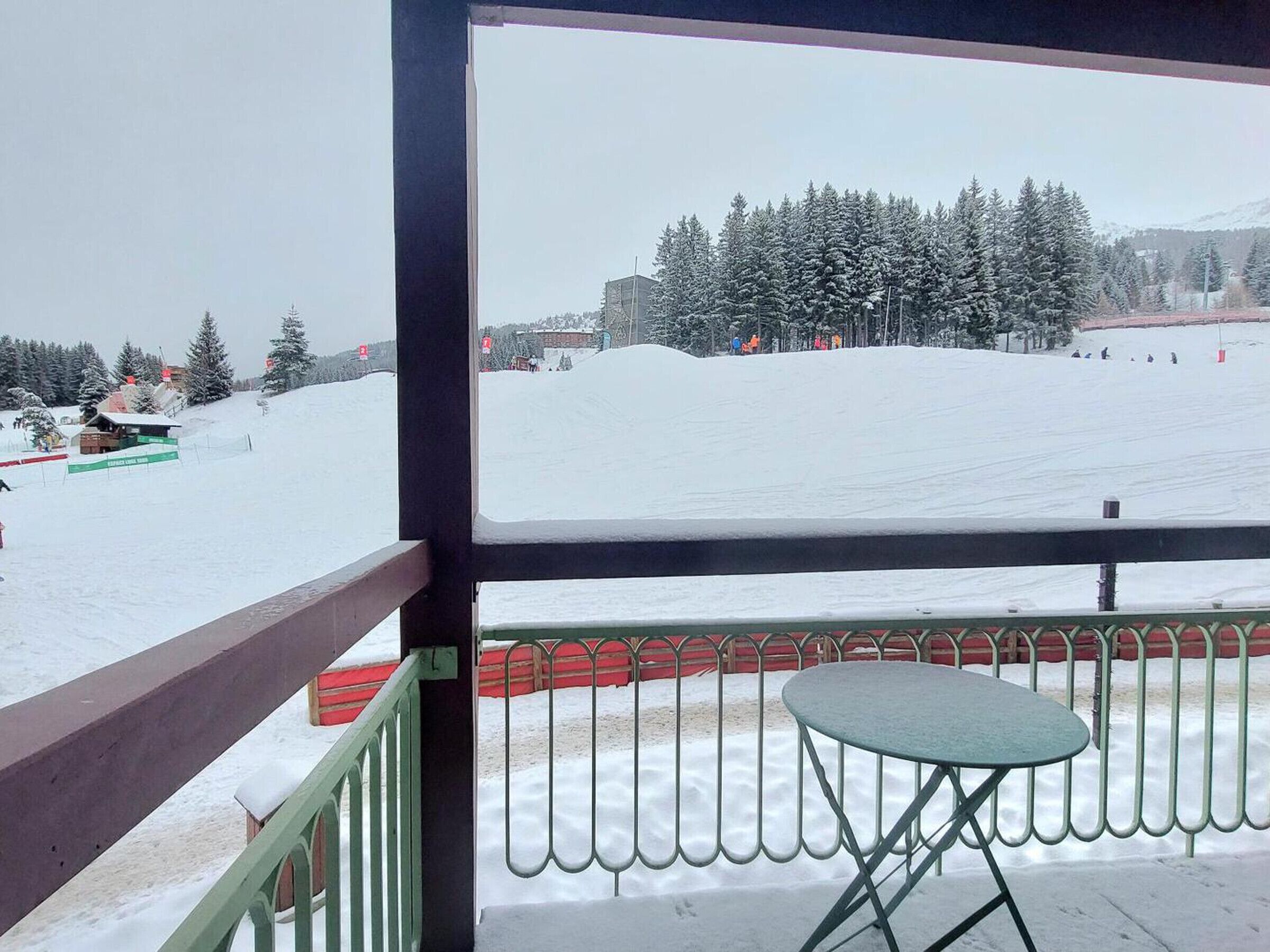 A view from a balcony with a metal railing and a small table, overlooking a snowy landscape with trees and a ski slope.
