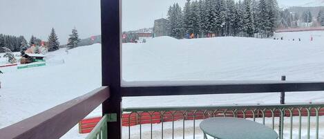 A view from a balcony with a metal railing and a small table, overlooking a snowy landscape with trees and a ski slope.