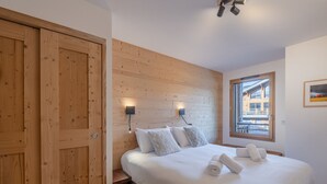 A bedroom with a wooden headboard, a large bed with white linens, a wooden bench, and a window with a view of buildings. - Modern apartment in Morzine, 6 ppl, Wi-Fi, parking, near slopes (Morzine)