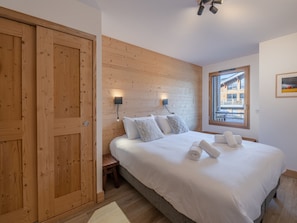 A bedroom with a wooden headboard, a large bed with white linens, a wooden bench, and a window with a view of buildings. - Modern apartment in Morzine, 6 ppl, Wi-Fi, parking, near slopes (Morzine)