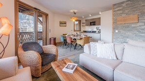 A modern living room with a sofa, a wicker chair, and a wooden coffee table. There is a dining area with a table and chairs, and a kitchen in the background. - Modern apartment in Morzine, 6 ppl, Wi-Fi, parking, near slopes (Morzine)