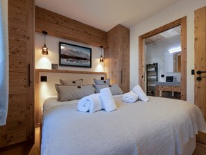 Ein Schlafzimmer mit hölzernem Kopfteil, einem Bett mit weißen Leinen, einem Gemälde von Bergen und einem durchgegangenen Türrahmen sichtbaren Badezimmer.
