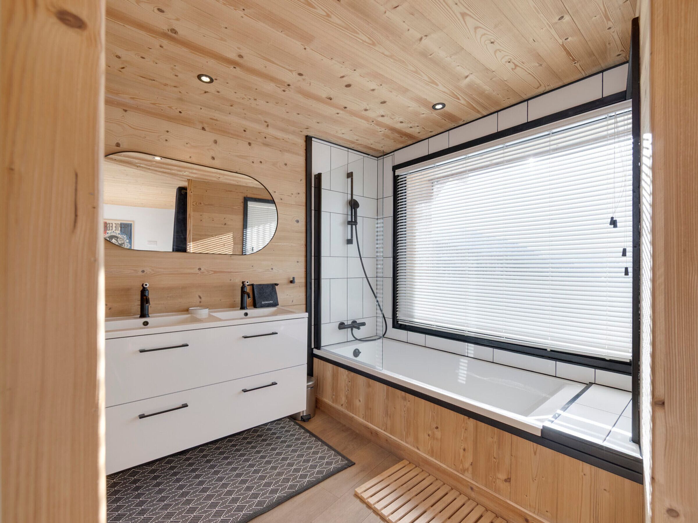 Une salle de bain moderne avec un plafond en bois, une grande fenêtre avec des stores, un miroir, un lavabo et un espace douche.