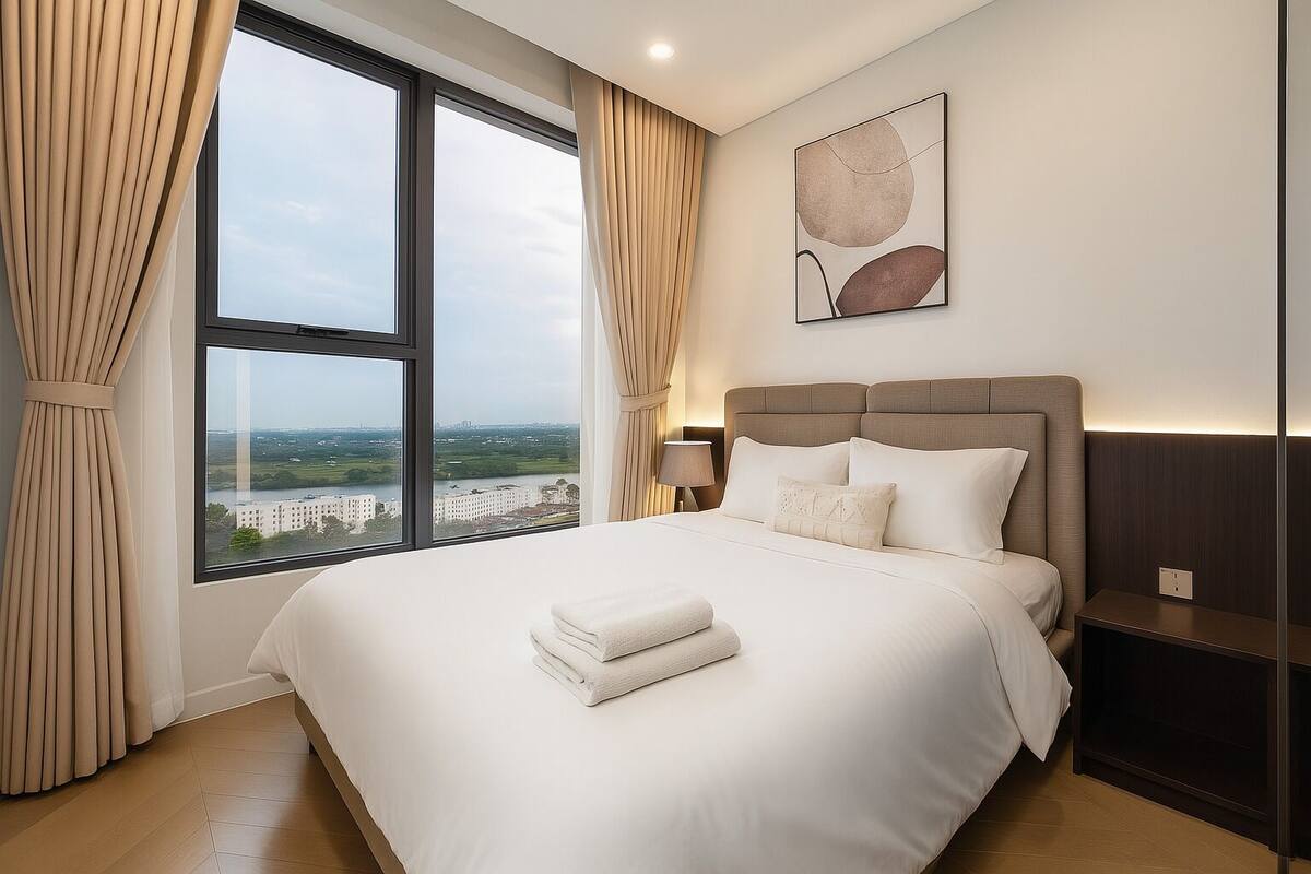 A hotel room with a large window, a bed with white linens, a headboard, a bedside table, and a wall-mounted lamp.