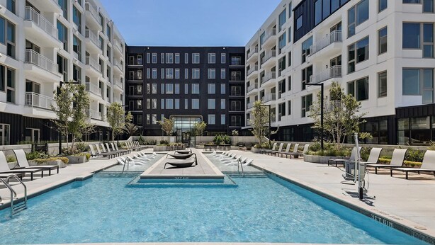 Un espace piscine moderne, doté de chaises longues et d’une fontaine centrale, entouré d’immeubles à plusieurs étages avec des balcons.