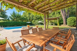 Eine Terrass mit Holzbank und Stühlen, einem Schwimmbad und einer Pergola mit Weinreben.