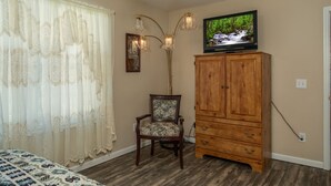 Ein Schlafzimmer mit einem Holzschrank, einem Fernseher darauf, einem blumigen Sessel und einem Bett mit gemusterten Bettbezügen.