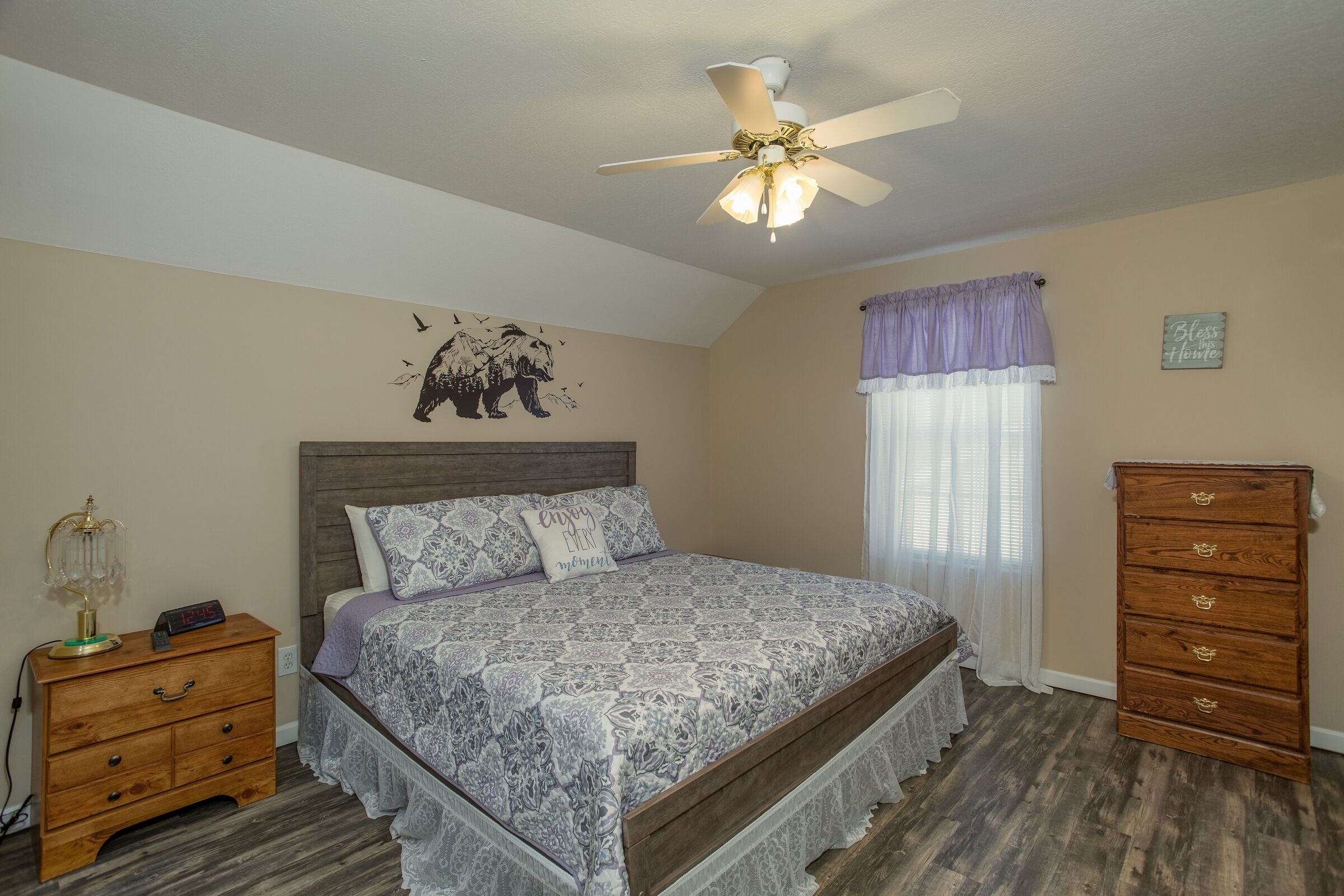 Une chambre à coucher comprenant un lit, une table de chevet, une commode, un ventilateur de plafond et un élément décoratif mural.