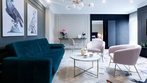 A modern living room with a teal sofa, a pink armchair, and a coffee table. There are framed artworks on the wall, a chandelier, and a view into another room. - Central Kraków 1-Unit Queen Bed Retreat | Breakfast Included (Kraków)