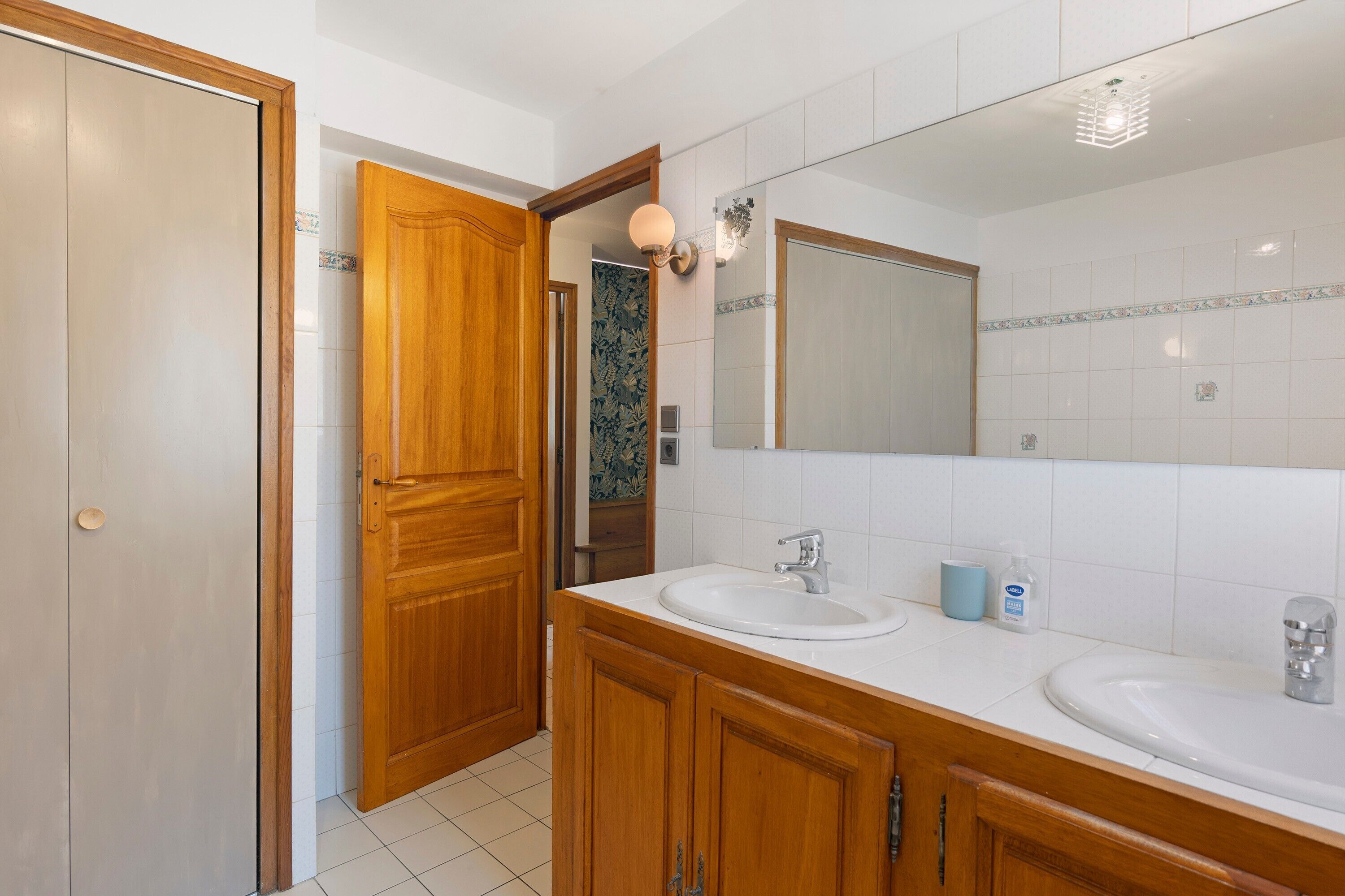A bathroom with two sinks, wooden cabinets, a large mirror, and a door leading to another room.