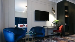 A modern hotel room with a large bed, two blue armchairs, a small table, a red chair, a TV, and a dark wooden bookshelf. - Central Kraków Classic King | Ideal for 2 | Free Bfast (Kraków)