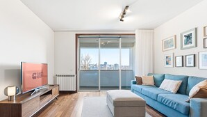 Ein modernes Wohnzimmer mit einem Flachbildfernseher, einer Holztrennwand, einem blauen Sofa und Blick auf die Stadt durch eine Schiebeglastür.