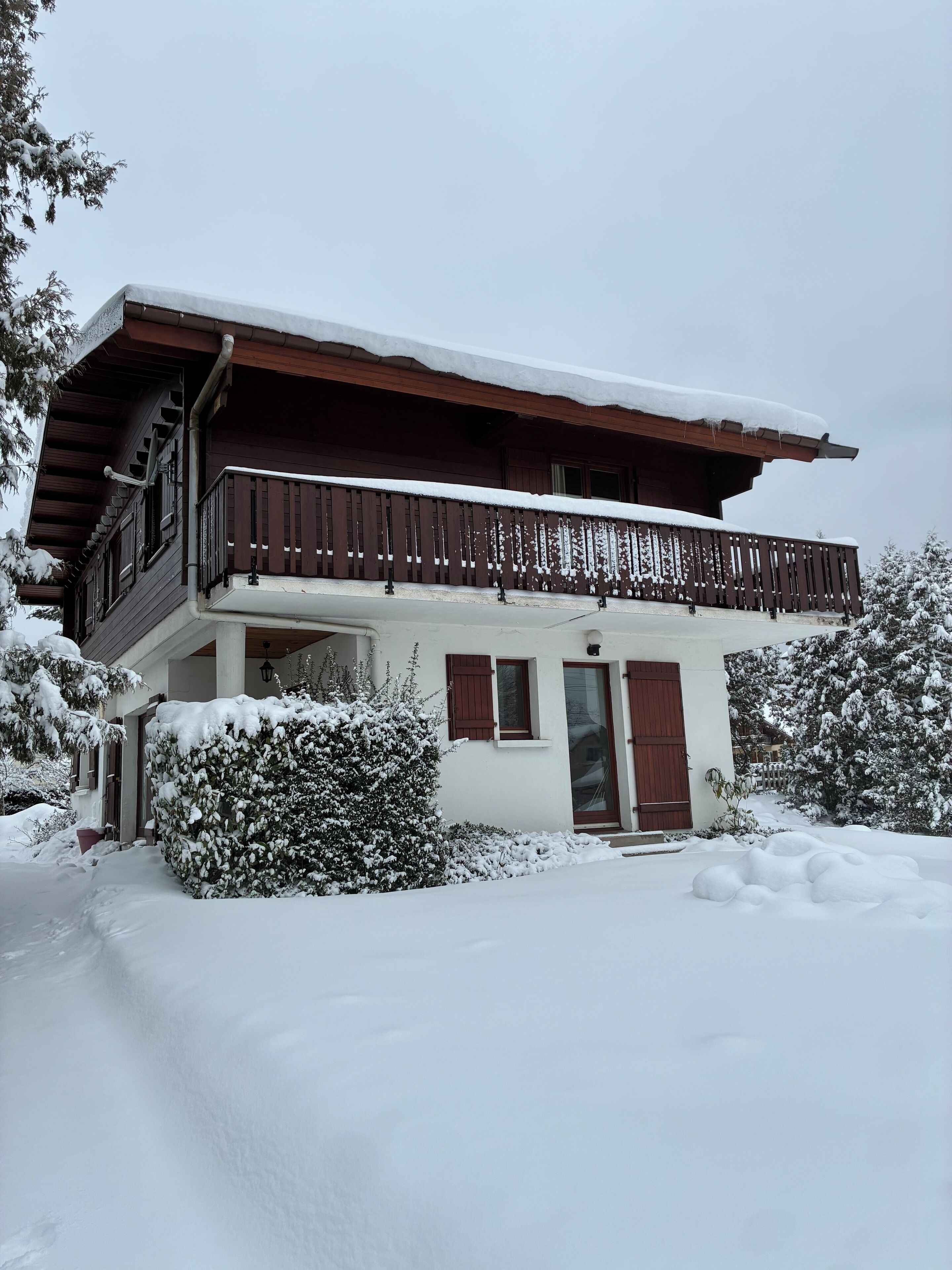 A two-story chalet with a wooden balcony, surrounded by snow-covered trees and bushes.