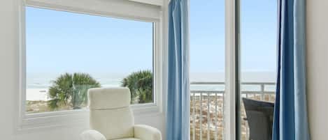 Ein Zimmer mit weißem Stuhl, einem großen Fenster mit Meerblick und einem Balkon mit blauen Vorhängen.