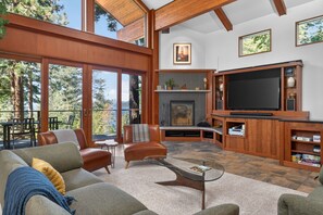 Ein großzügiges Wohnzimmer mit Kamin, einem großen Flachbildfernseher und einem Balkon mit Blick auf Bäume.