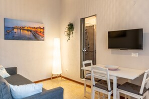 A modern room with a grey sofa, a white table with chairs, a TV, and a painting of a cityscape. - Casa le Radici by Wonderful Italy (Syracuse)
