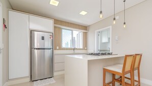 A modern kitchen with a stainless steel refrigerator, a central island, and wooden chairs. - Apartamento no Centro com 3 su tes a 400m da Praia (Balneário Camboriú)