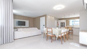 A modern, well-lit living area with a white sofa, dining table, and chairs. A flat-screen TV is mounted on the wall, and a kitchen area is visible in the background. - Apartamento no Centro com 3 su tes a 400m da Praia (Balneário Camboriú)