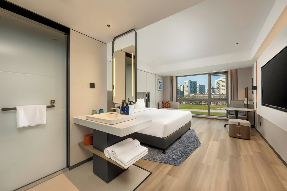 Una habitación de hotel moderna con una cama grande, un baño con tocador, un espejo amplio y vistas de la ciudad a través de las ventanas.