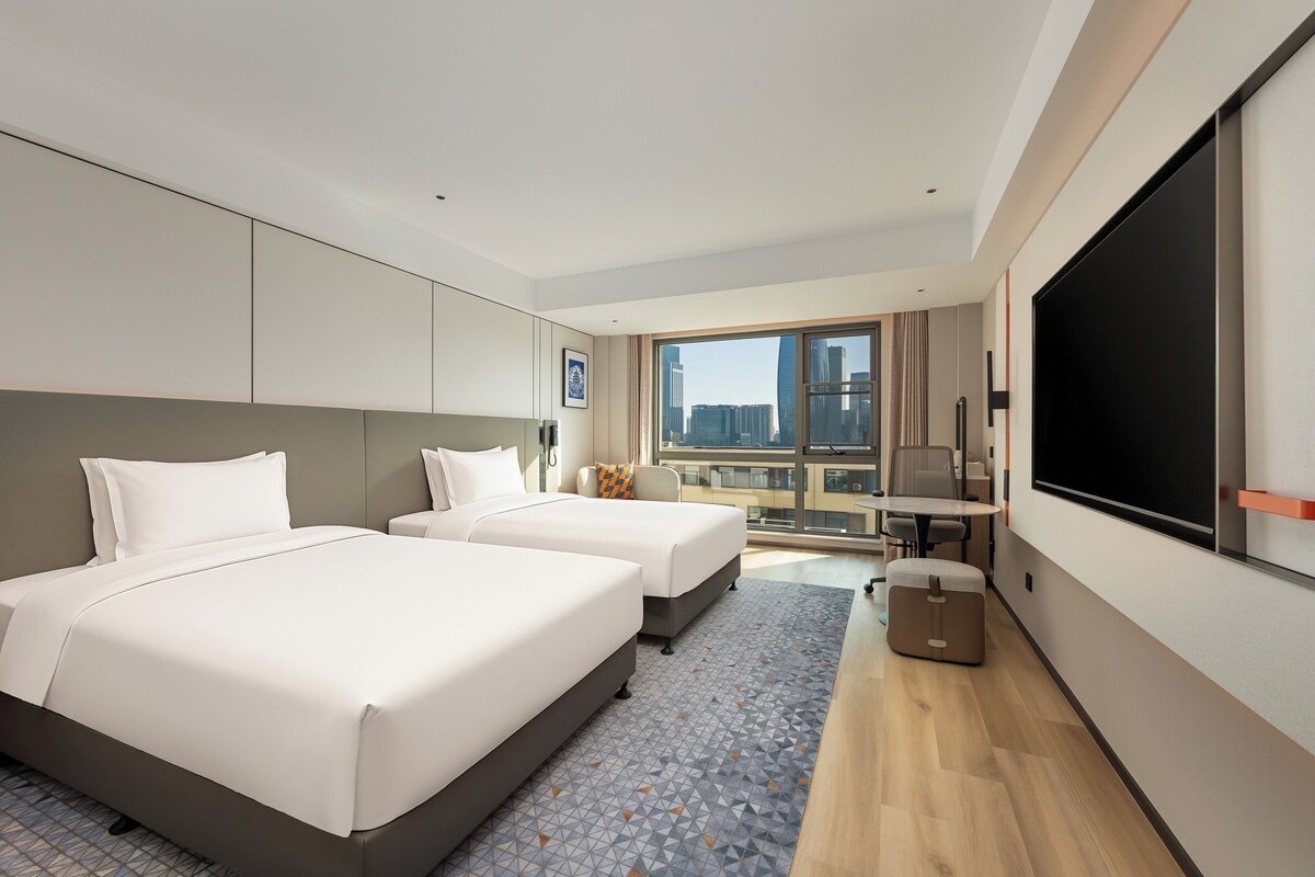Een hotelkamer met twee bedden, een bureau, een stoel, een televisie en uitzicht op de stad.
