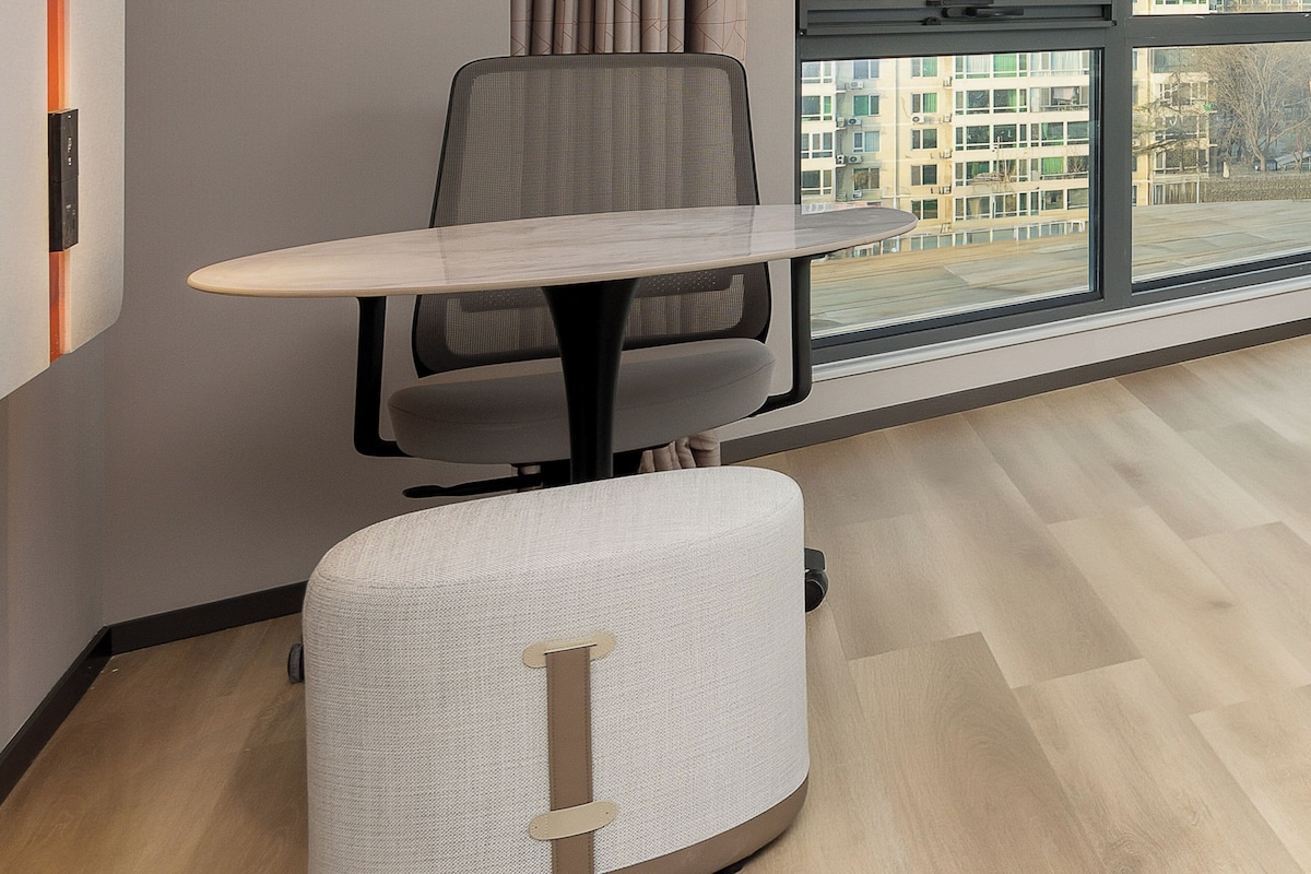 A modern office space with a round table, a single office chair, and a large window offering a view of buildings.