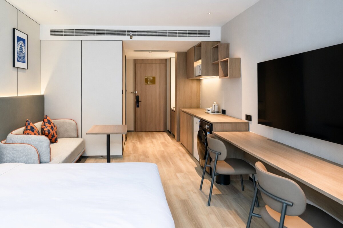 Een moderne hotelkamer met een bed, een bureau, een stoel, een televisie, een kitchenette en een deur naar een badkamer.