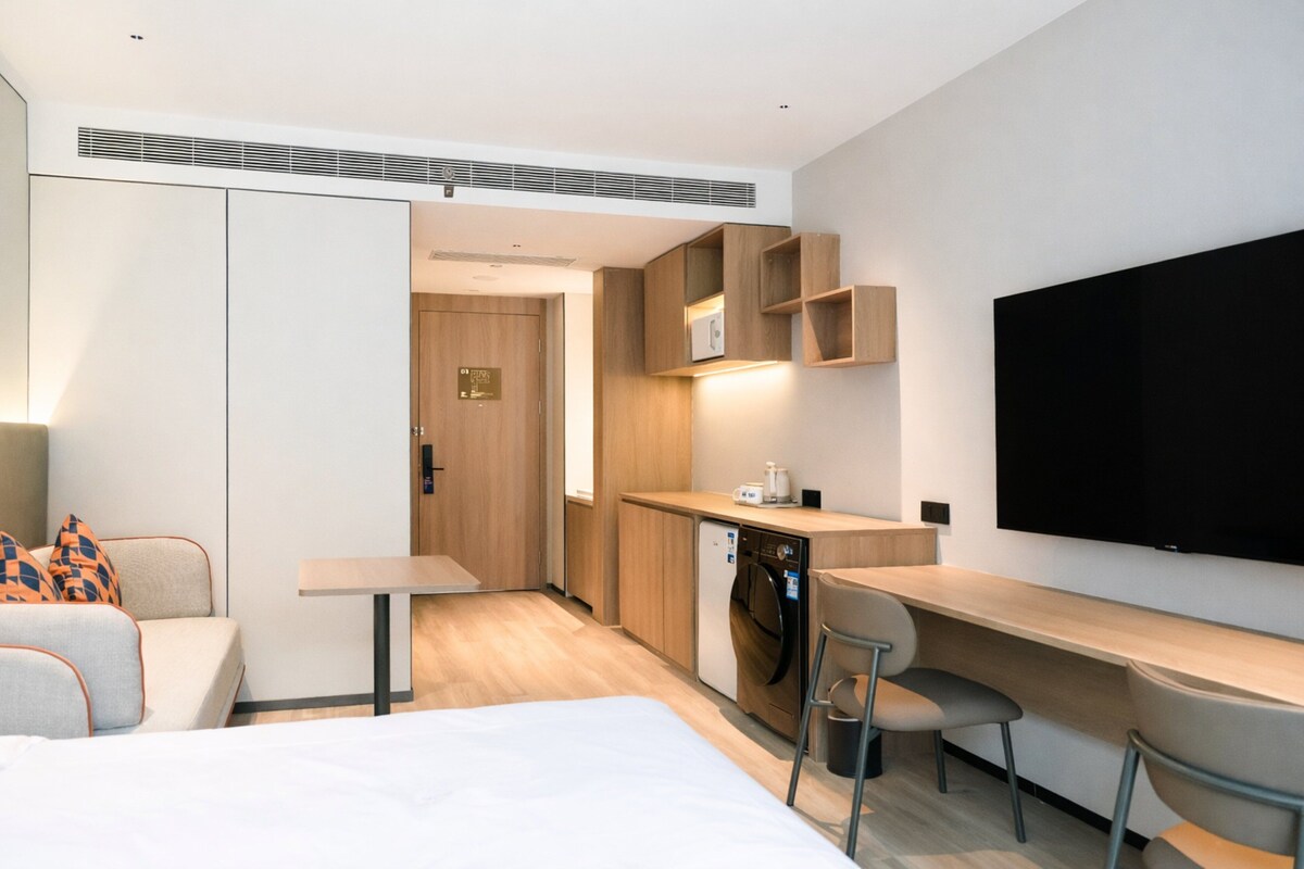 Una habitación de hotel moderna con cama, escritorio, silla, una kitchenette pequeña y televisión montada en la pared.