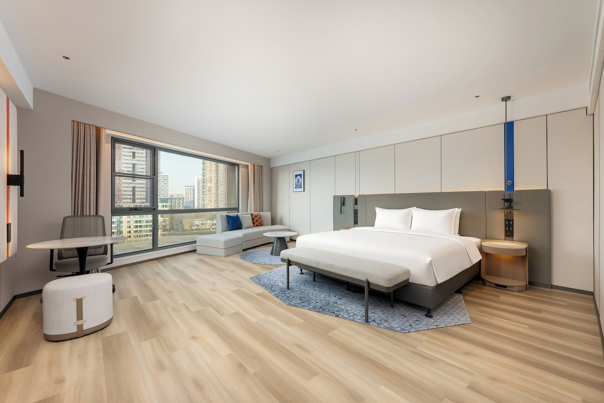 A modern hotel room with a large bed, a desk, a sofa, and a view of the city.