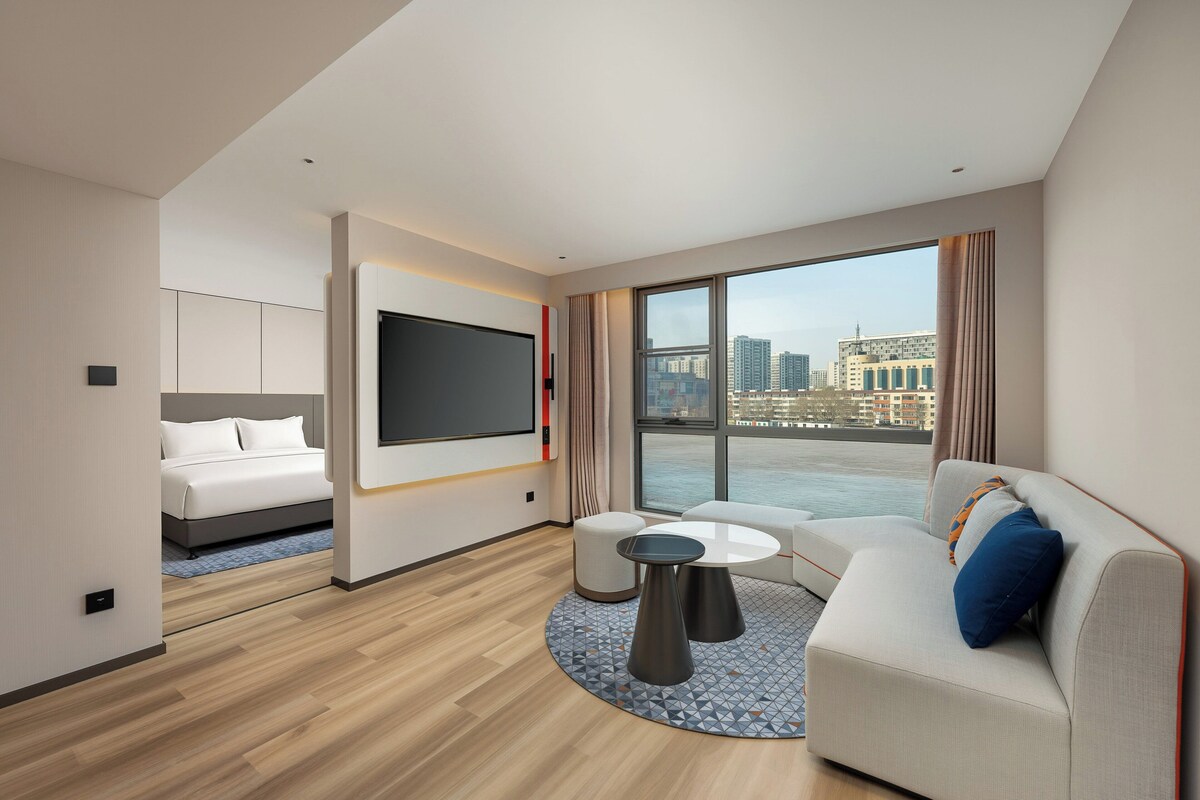 Een moderne hotelkamer met een bed, een televisie, een bank en een groot raam met uitzicht op de stad.