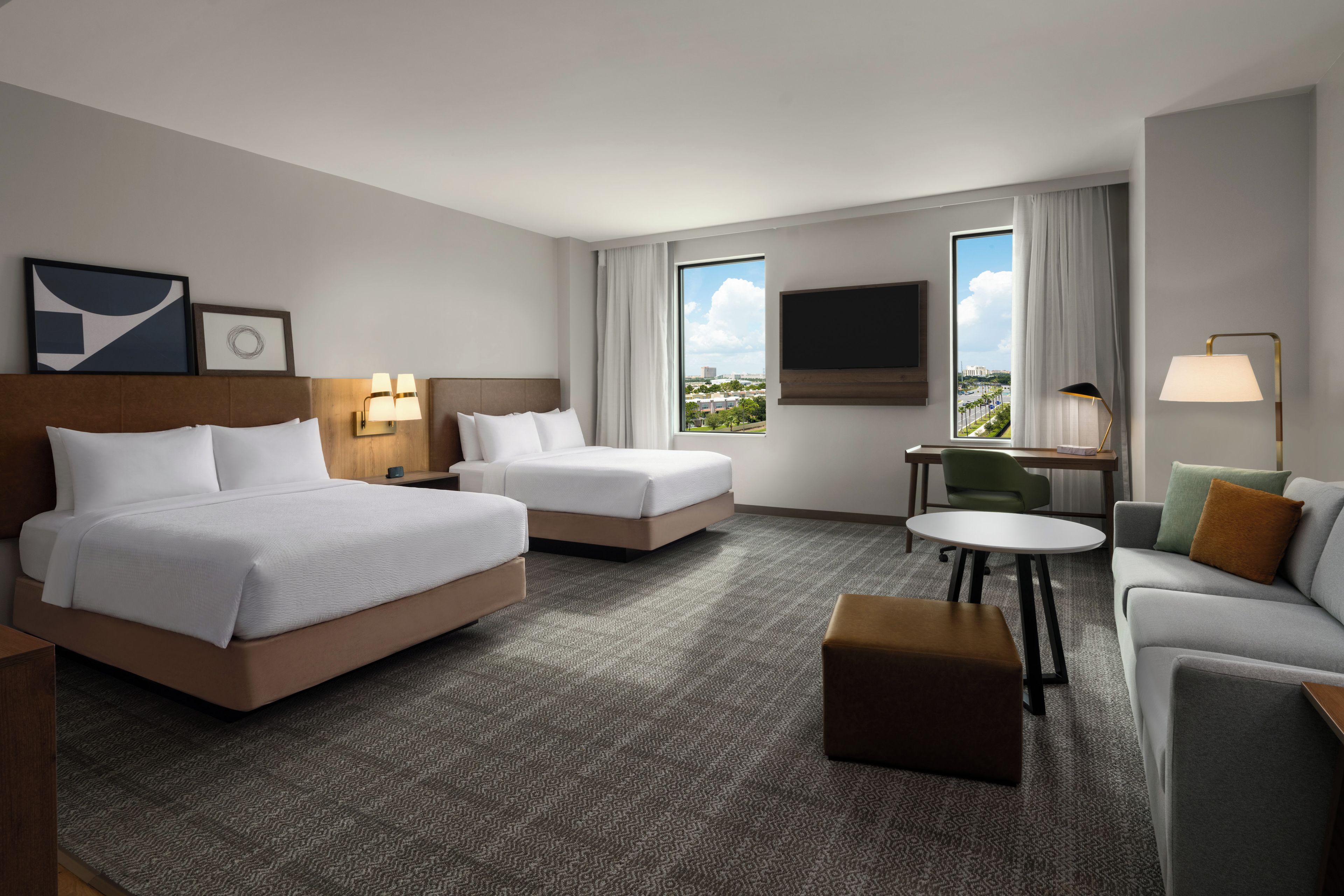 A hotel room with two beds, a sofa, a small table, a TV, and a window with a city view.