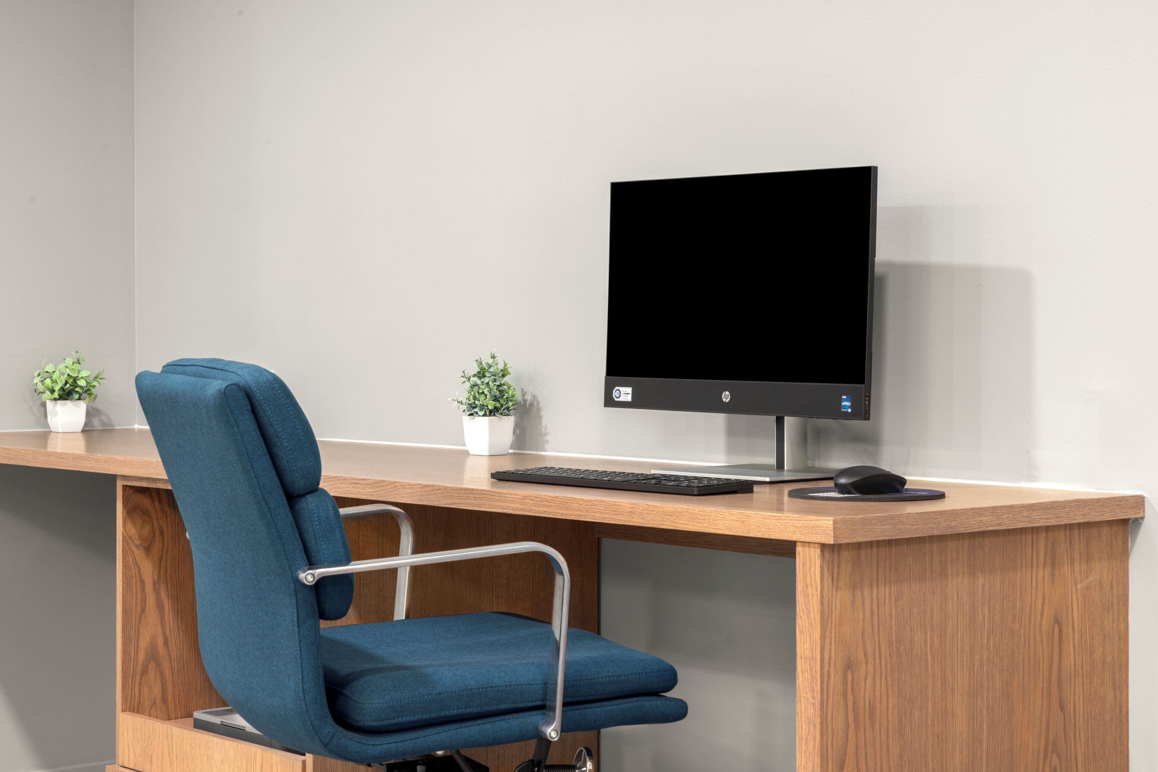 Un bureau moderne avec une chaise bleue, un écran d’ordinateur, un clavier et une souris. Deux petites plantes en pot sont posées sur le bureau.
