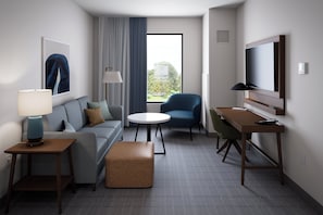 A modern hotel room with a sofa, a round table, a blue armchair, a wooden desk, a TV mounted on the wall, and a large window with a view of greenery. - Staybridge Suites Orlando Universal Blvd by IHG (Orlando)