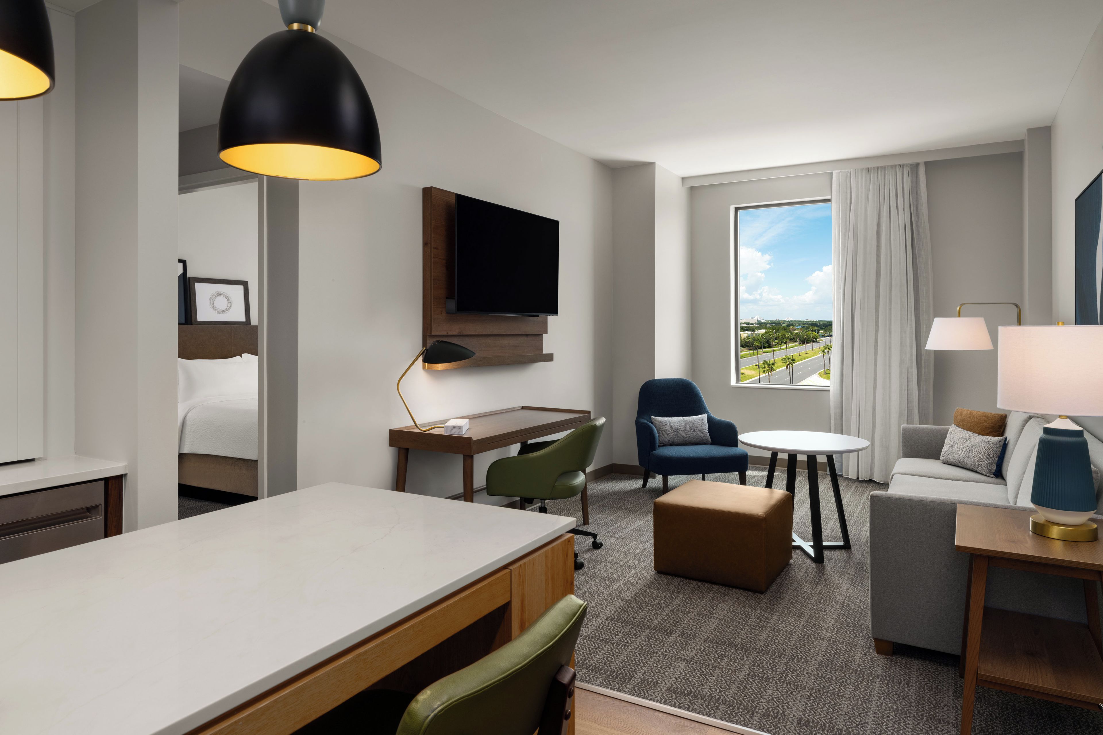 A hotel room with a flat-screen TV, a desk, a dining table, a sofa, a chair, and a window with a view of a road and greenery.