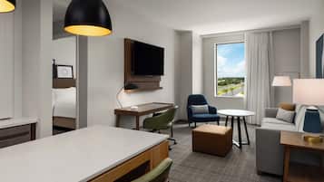 A hotel room with a flat-screen TV, a desk, a dining table, a sofa, a chair, and a window with a view of a road and greenery.