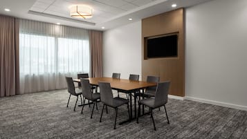 A conference room with a wooden table, six chairs, a flat-screen TV mounted on the wall, and a large window with curtains.
