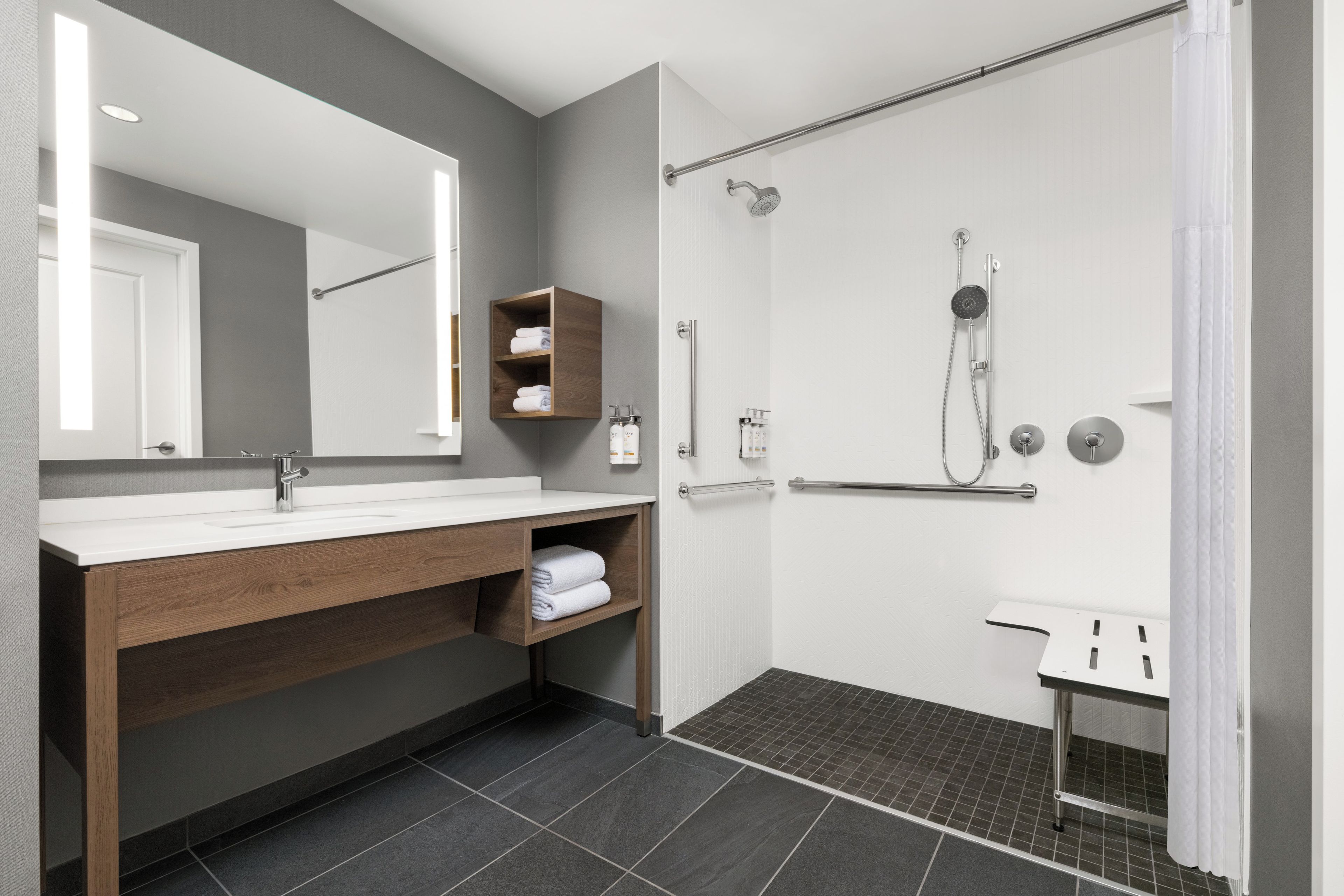 Une salle de bain moderne avec un grand miroir, un meuble-lavabo en bois, une douche équipée d’un pommeau de douche à main et un lavabo blanc.