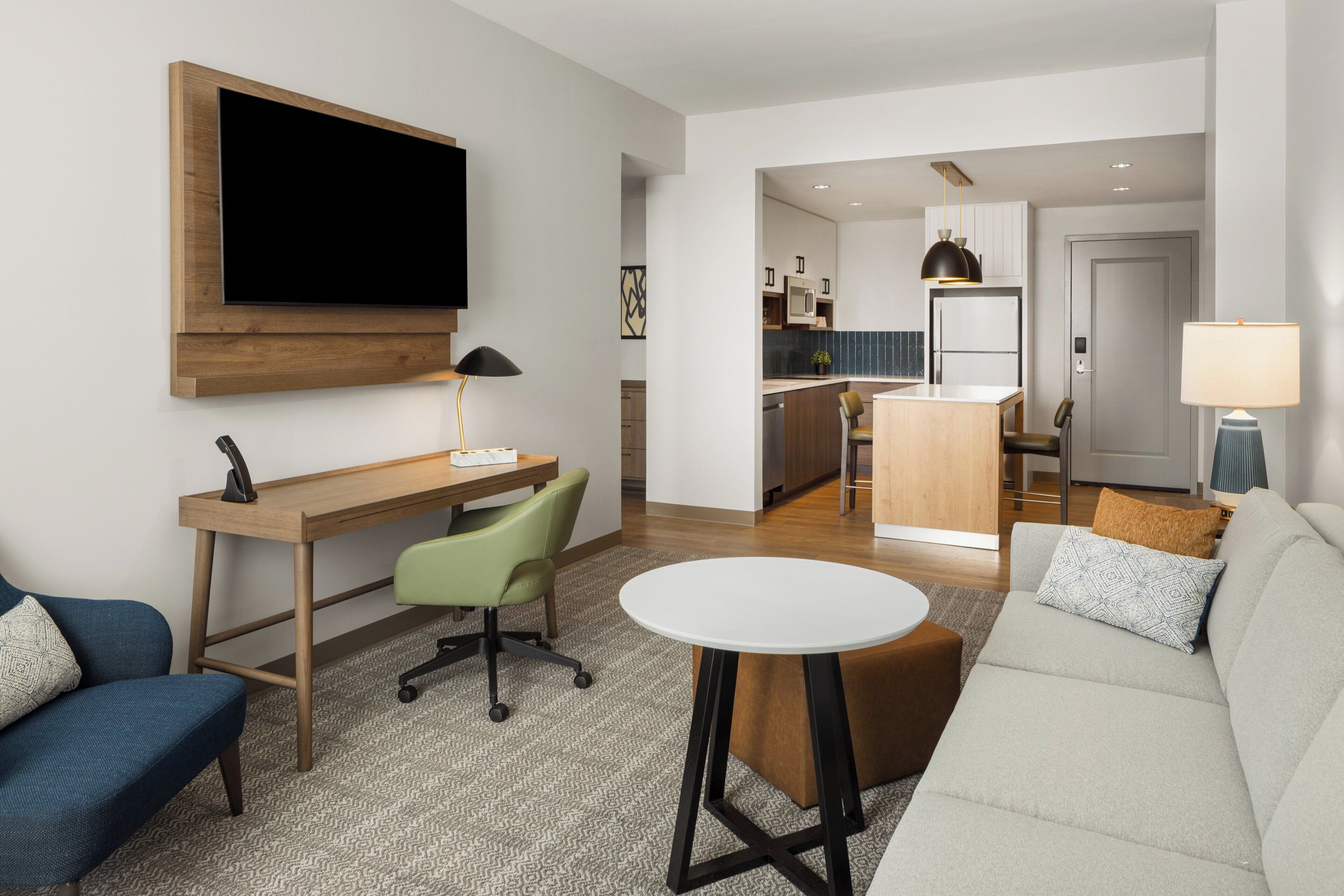 Een moderne woonkamer met een bank, een bureau, een stoel, een televisie en een keuken op de achtergrond.