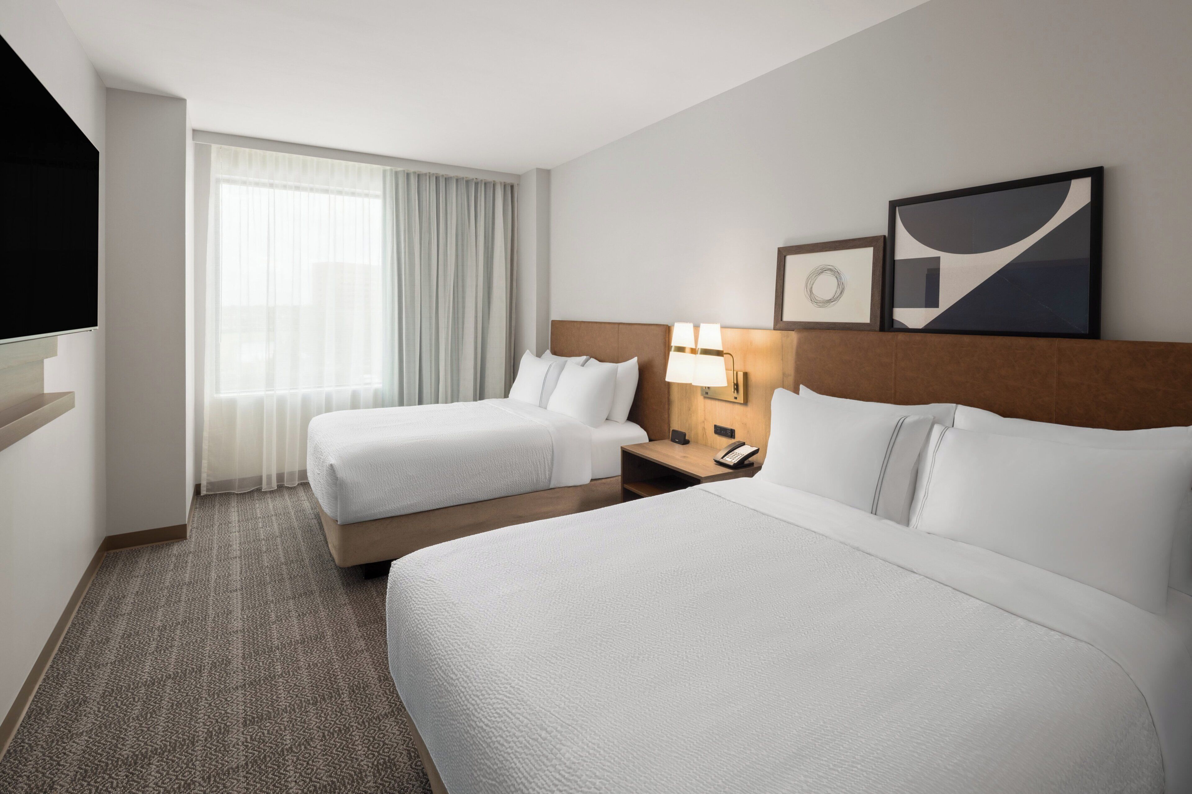 A hotel room with two beds, a nightstand, a lamp, and a television.