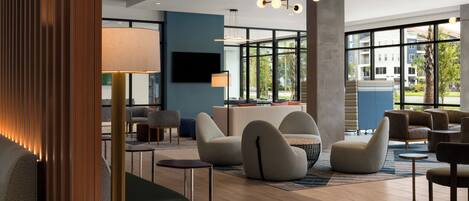 A modern hotel lobby with a seating area, a flat-screen TV, and large windows offering a view of the outside.
