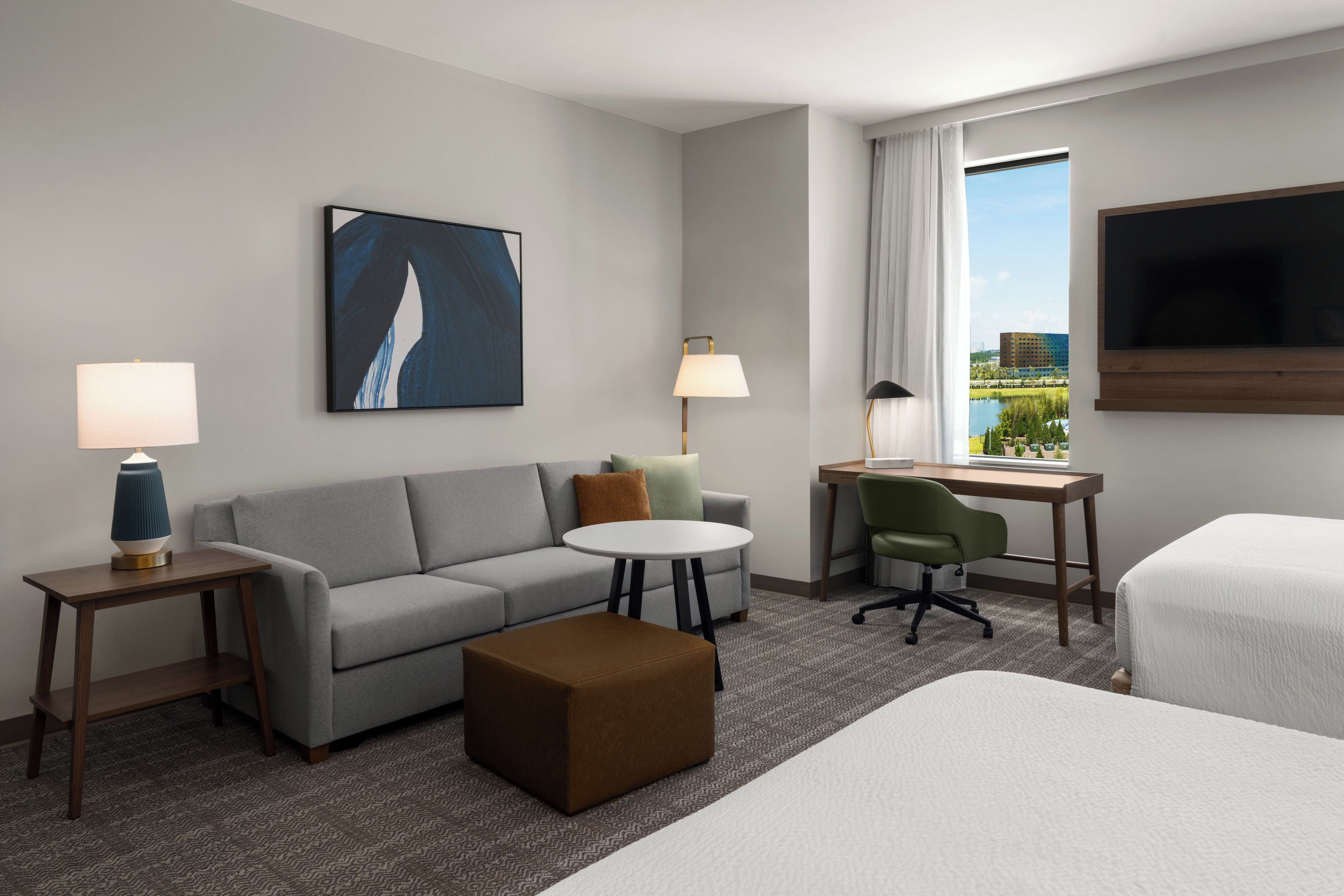 Une chambre d’hôtel avec un canapé gris, une petite table ronde, une chaise verte, un lit et un bureau avec un ordinateur.