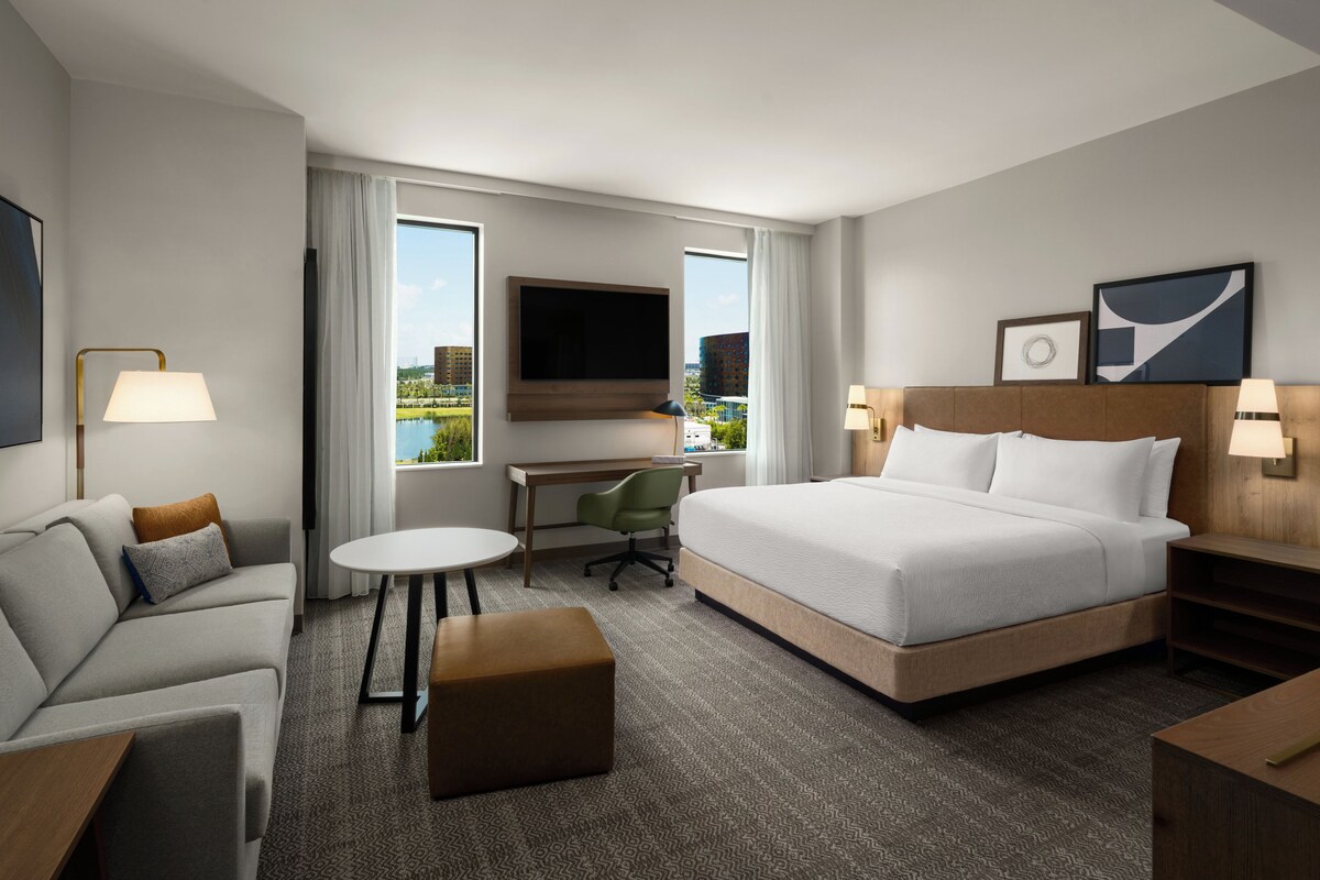 Habitación de hotel con cama, escritorio, silla, televisor y vista a la ciudad.