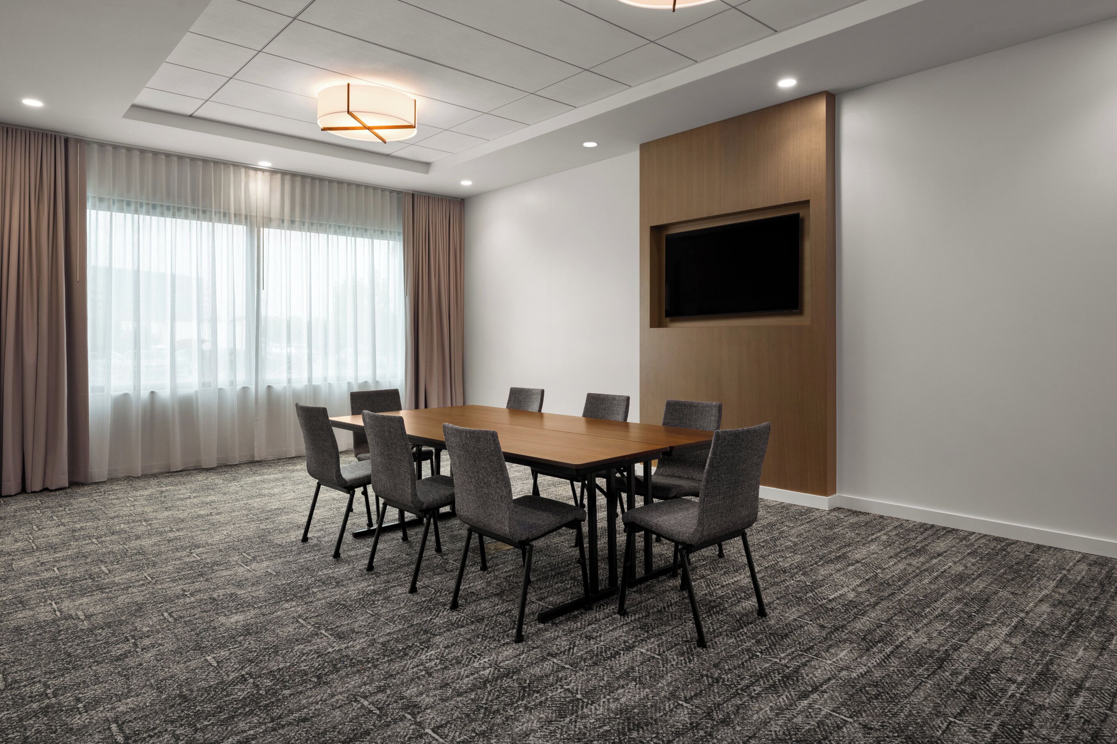 A conference room with a large table, six chairs, a flat-screen TV mounted on a wooden panel, and a ceiling with recessed lighting.