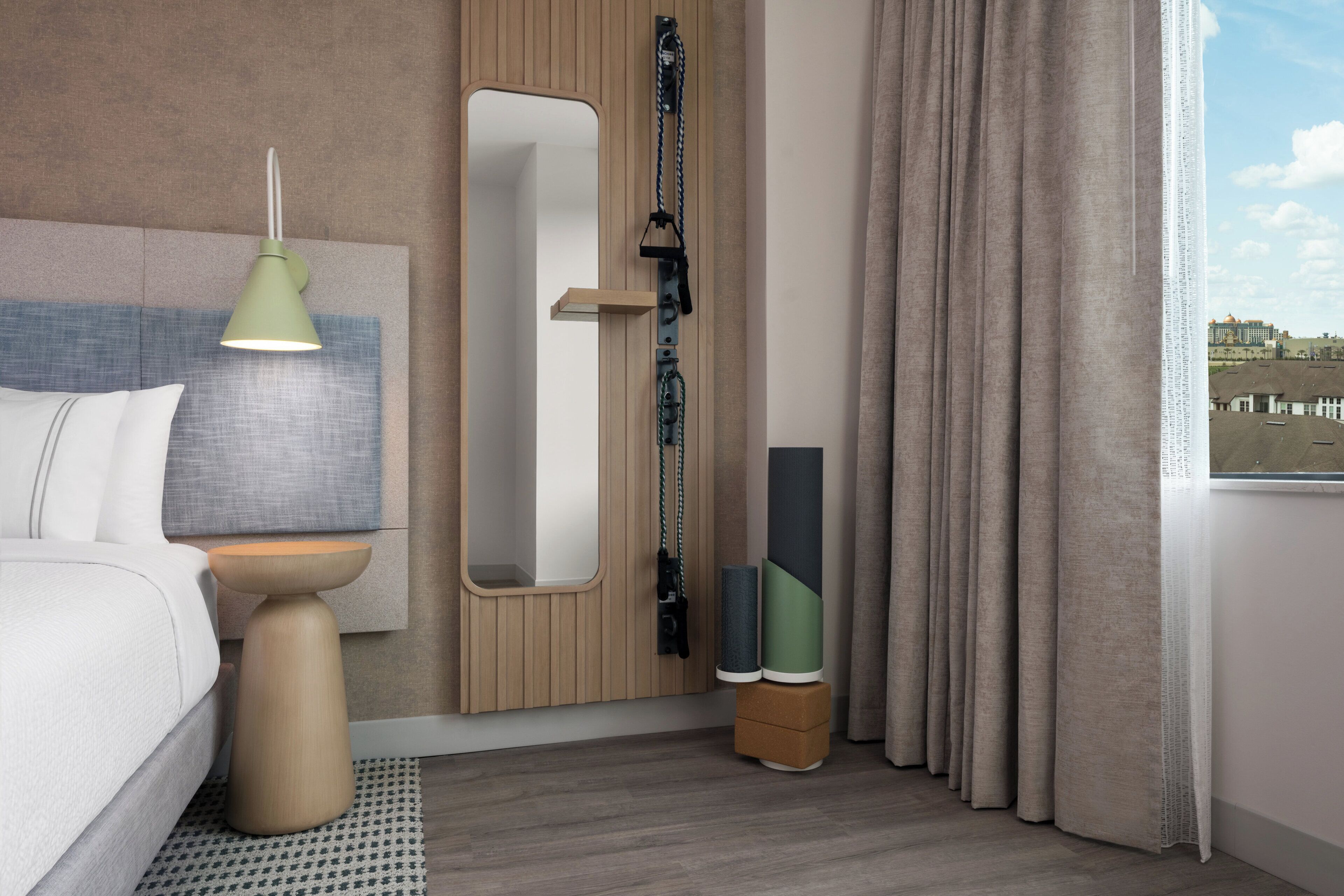 Una camera d'albergo con un letto, una lampada da comodino, uno specchio, una finestra con tende e vista su un edificio.