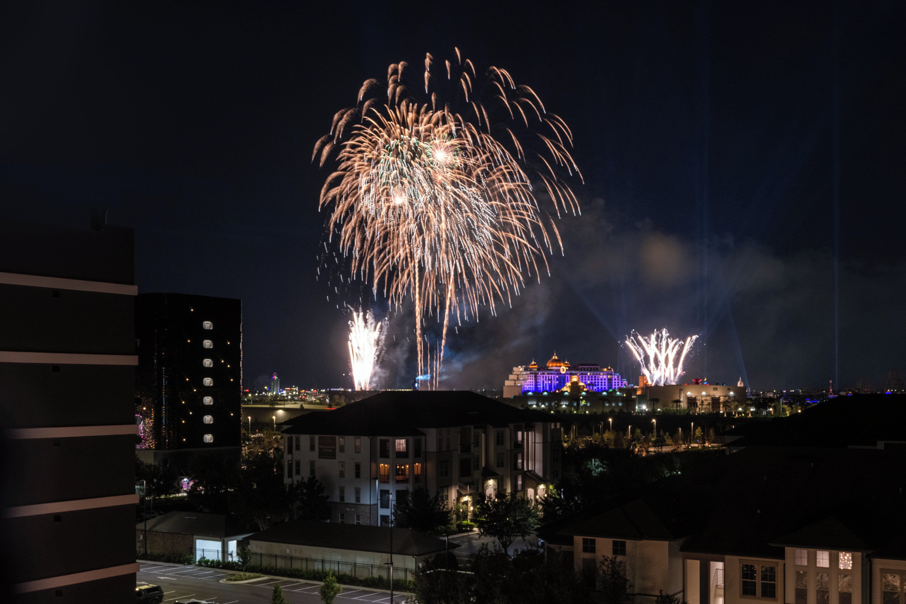 A nighttime view of a cityscape with fireworks in the sky, buildings with illuminated facades, and a parking area.