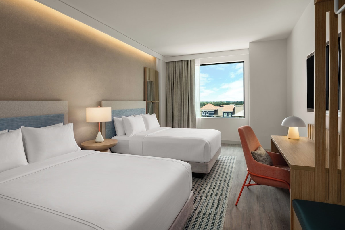 A hotel room with two beds, a desk, a chair, a TV, and a window with a view of buildings.
