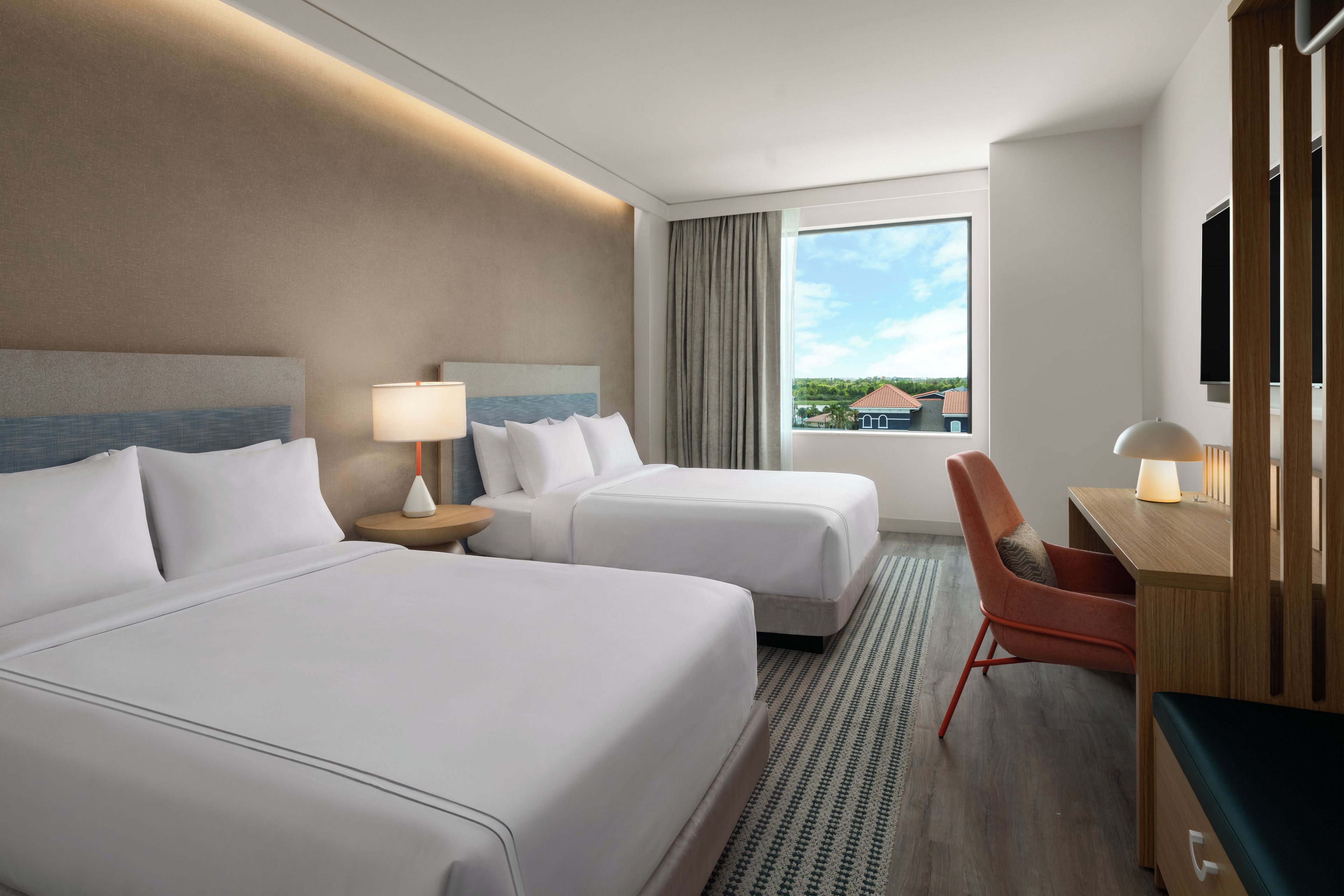 Habitación de hotel con dos camas, un escritorio, una silla, un televisor y ventana con vista a casas.