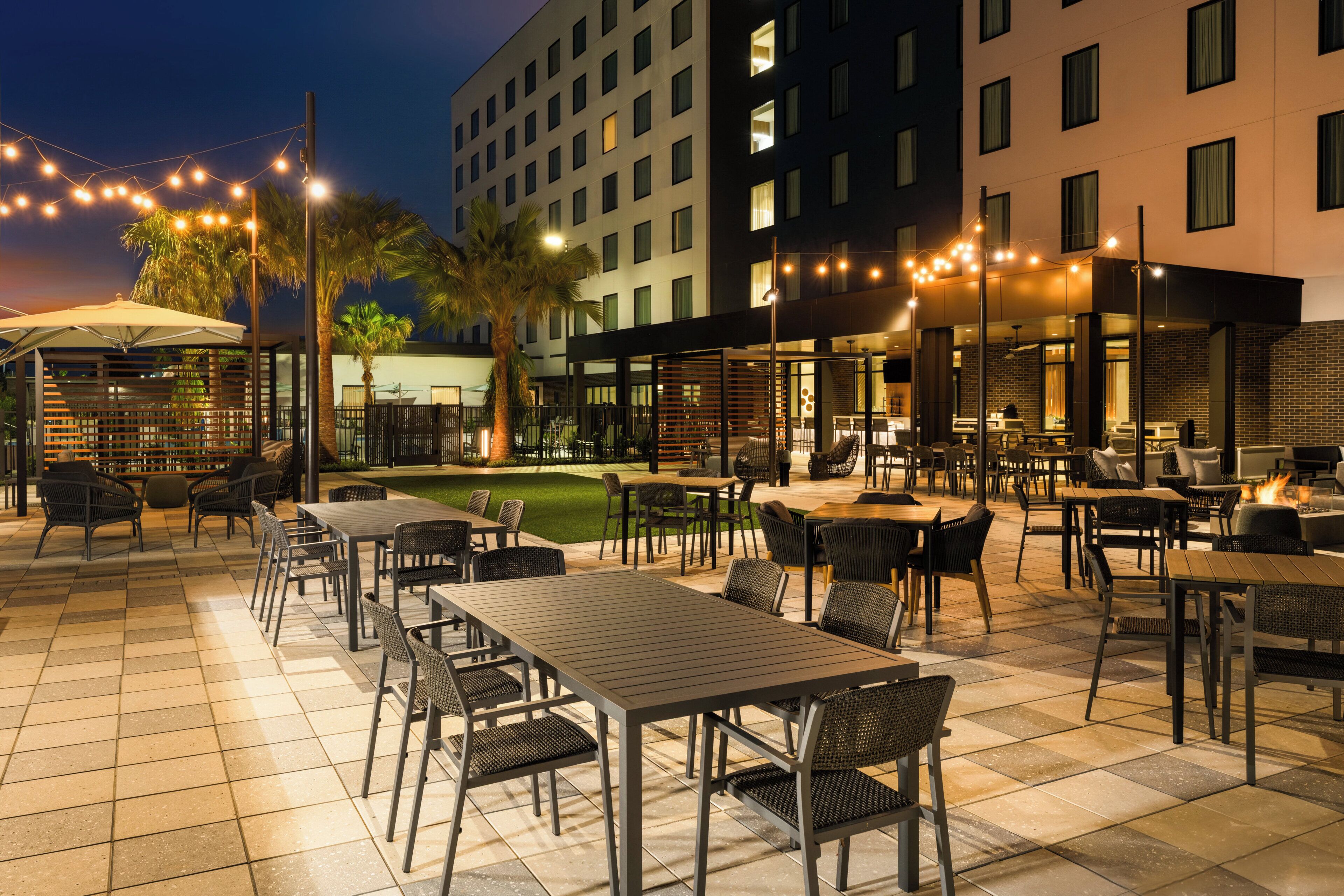 Un área de comedor al aire libre con mesas y sillas, rodeada por un edificio con varias ventanas.